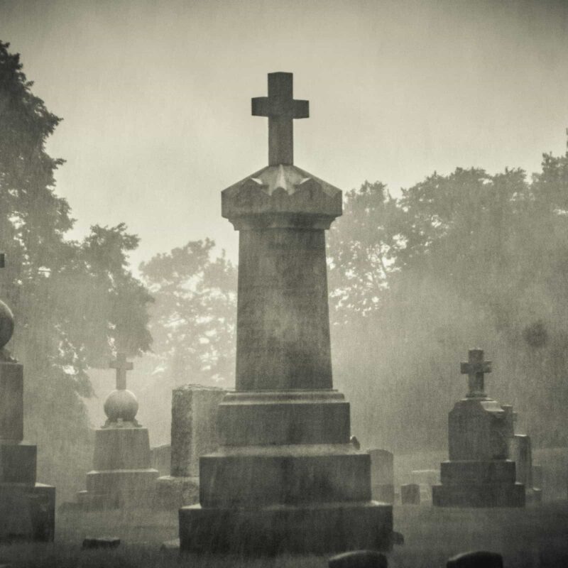 An dystopian looking scene of gravestones in a cemetery. The photo is in monochrome and it is raining.