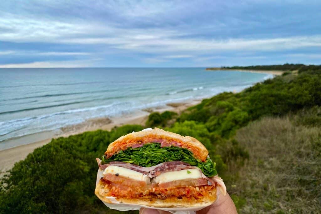A hand holding a delicious sandwich with the ocean in the background.
